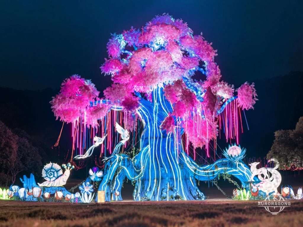 Giant illuminated LED tree landscape sculpture with pink and blue canopy, white birds, and flowers at night