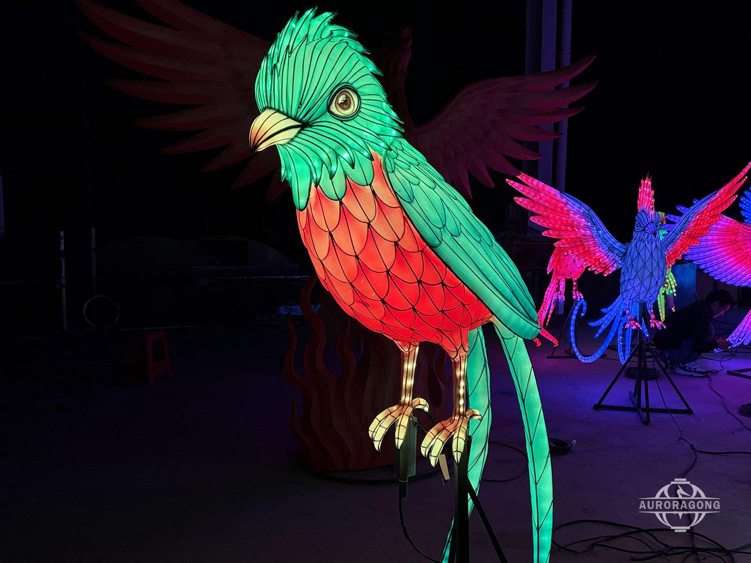 Large illuminated tropical bird lantern with green and red plumage and a long flowing tail