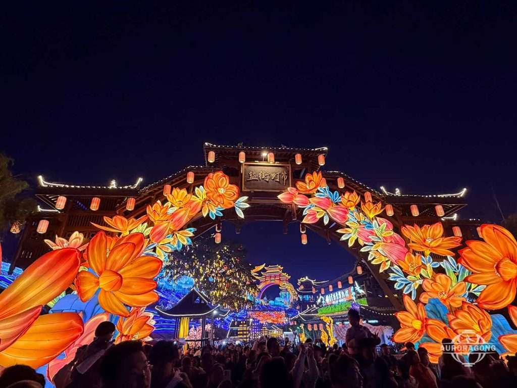 Nighttime festival entrance with a Chinese gate arch illuminated by colorful floral lanterns and festival crowds