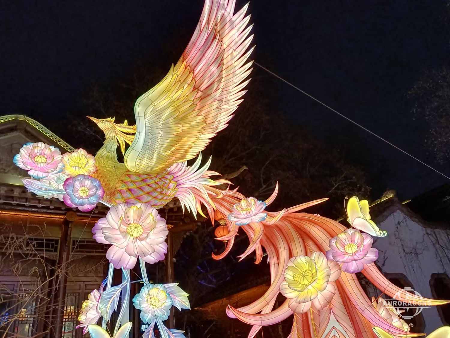 Side view of illuminated phoenix lantern with long tail feathers and flowers at night