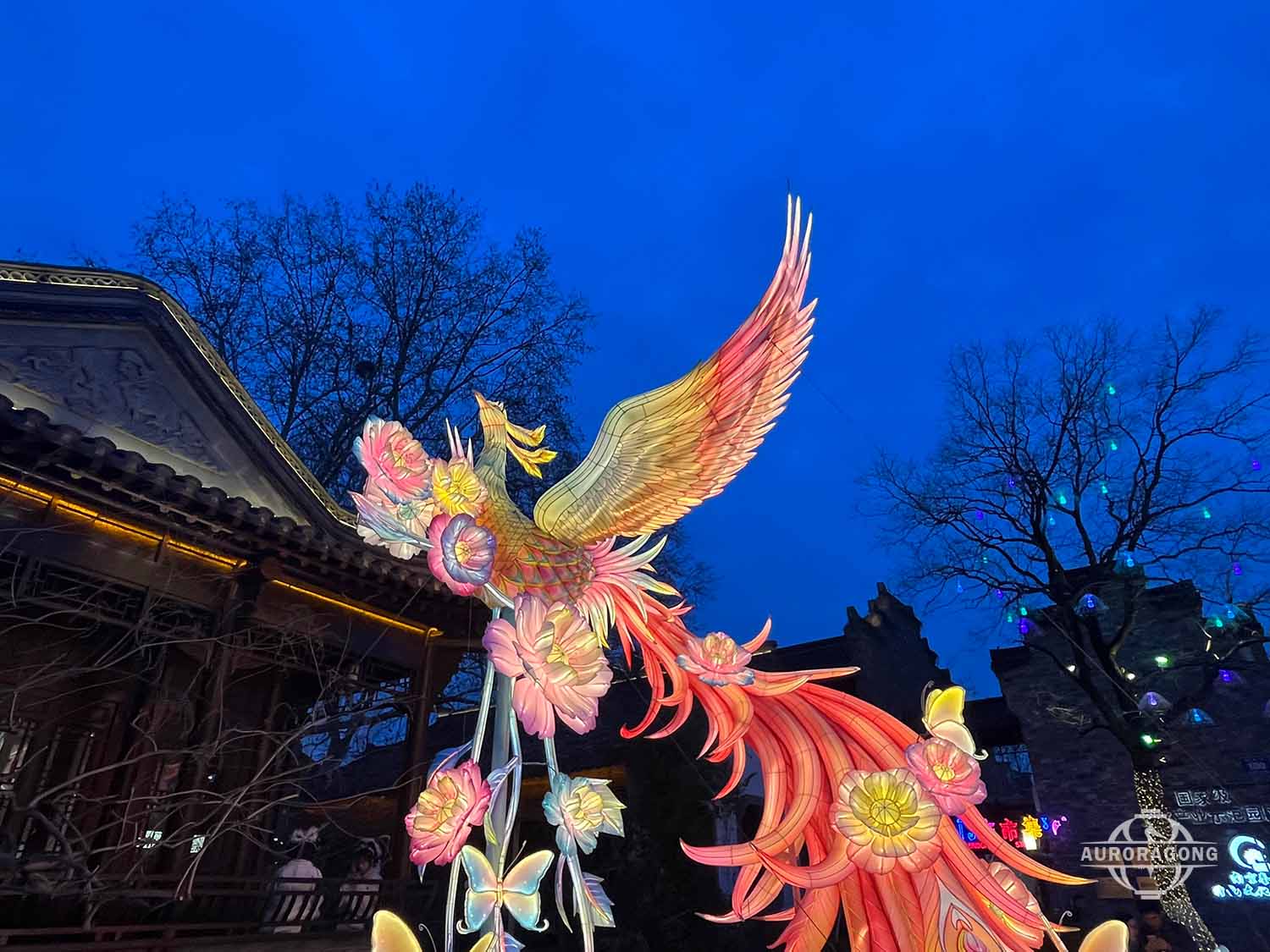 Custom illuminated phoenix lantern with yellow flowers and orange tail against blue dusk sky with traditional Chinese pavilion