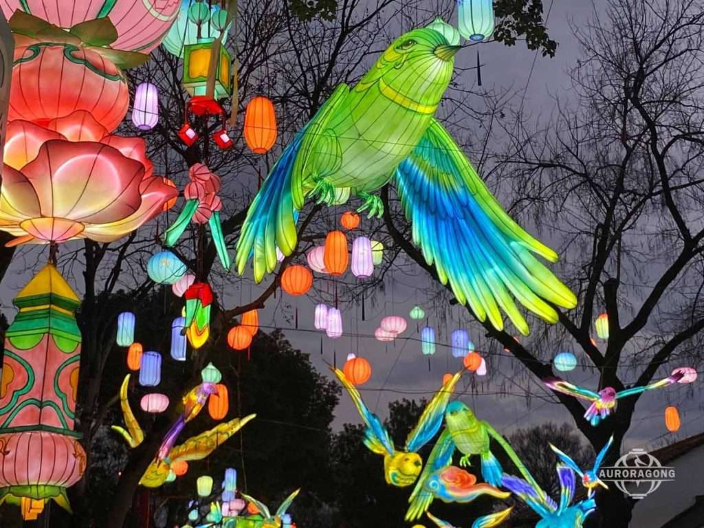 Large illuminated green parrot lantern hanging among colorful smaller lanterns in trees at dusk