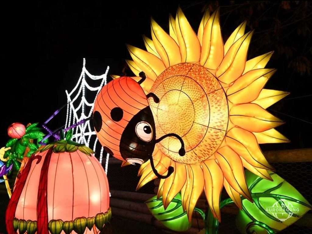Custom illuminated giant sunflower lantern with ladybug, spider web, and pink beetle at a garden evening event
