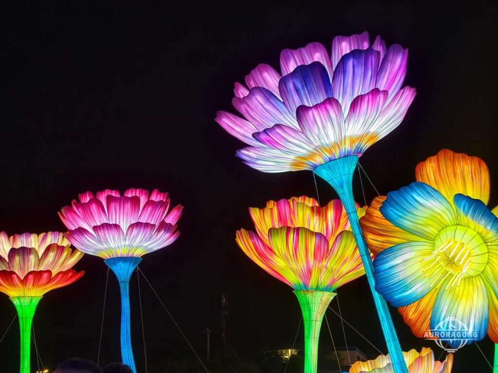 Tall illuminated flower lanterns in pink, purple, blue, and green against a dark night sky