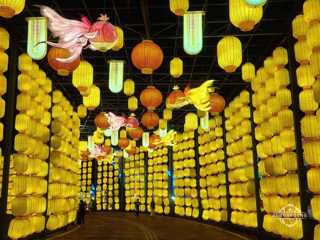 Long tunnel of yellow illuminated Chinese lanterns with pink fish lanterns hanging overhead