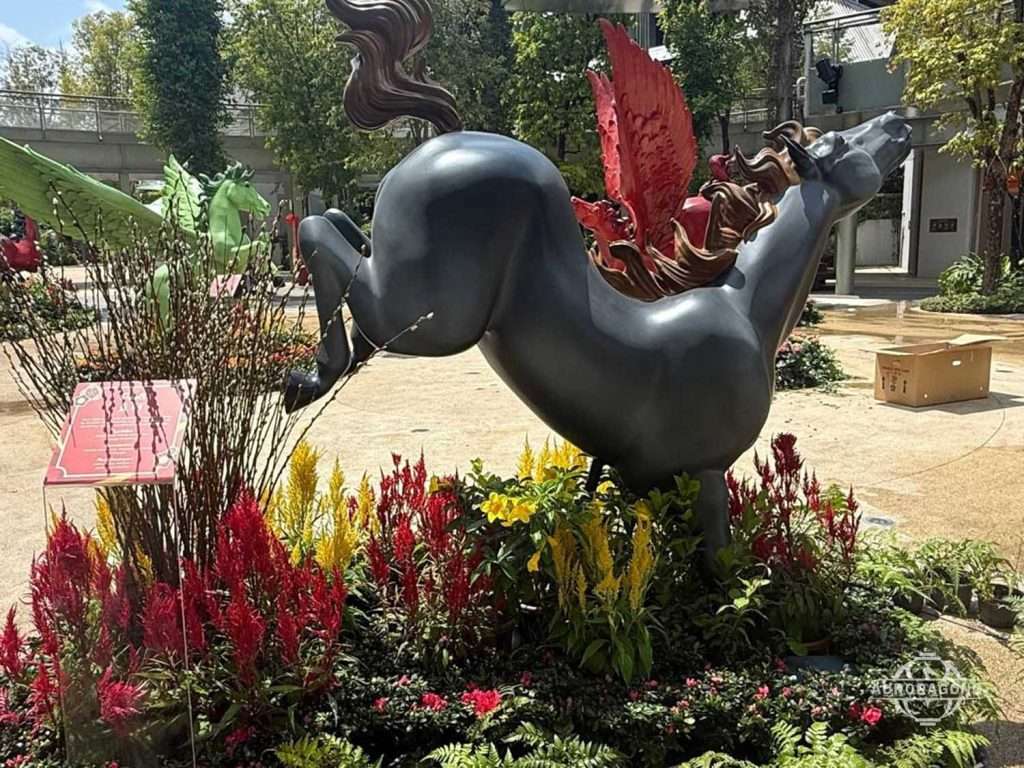 Black fiberglass horse sculpture in a rearing pose displayed among red flowers in an outdoor plaza