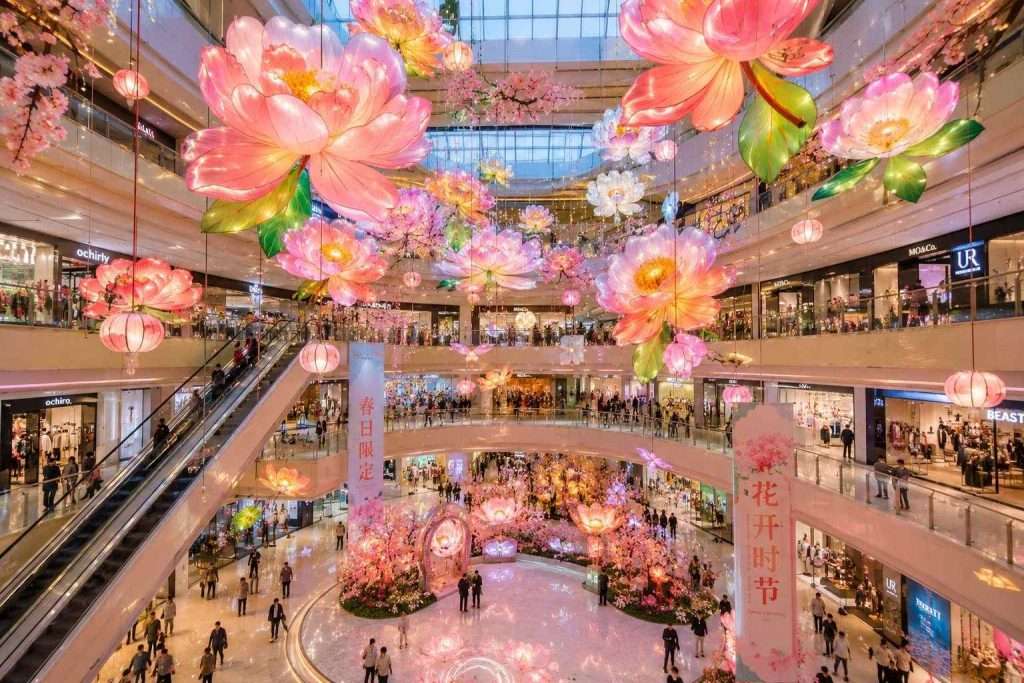 Pink peony hanging lantern installation decorating a multi-floor shopping mall atrium with shoppers below