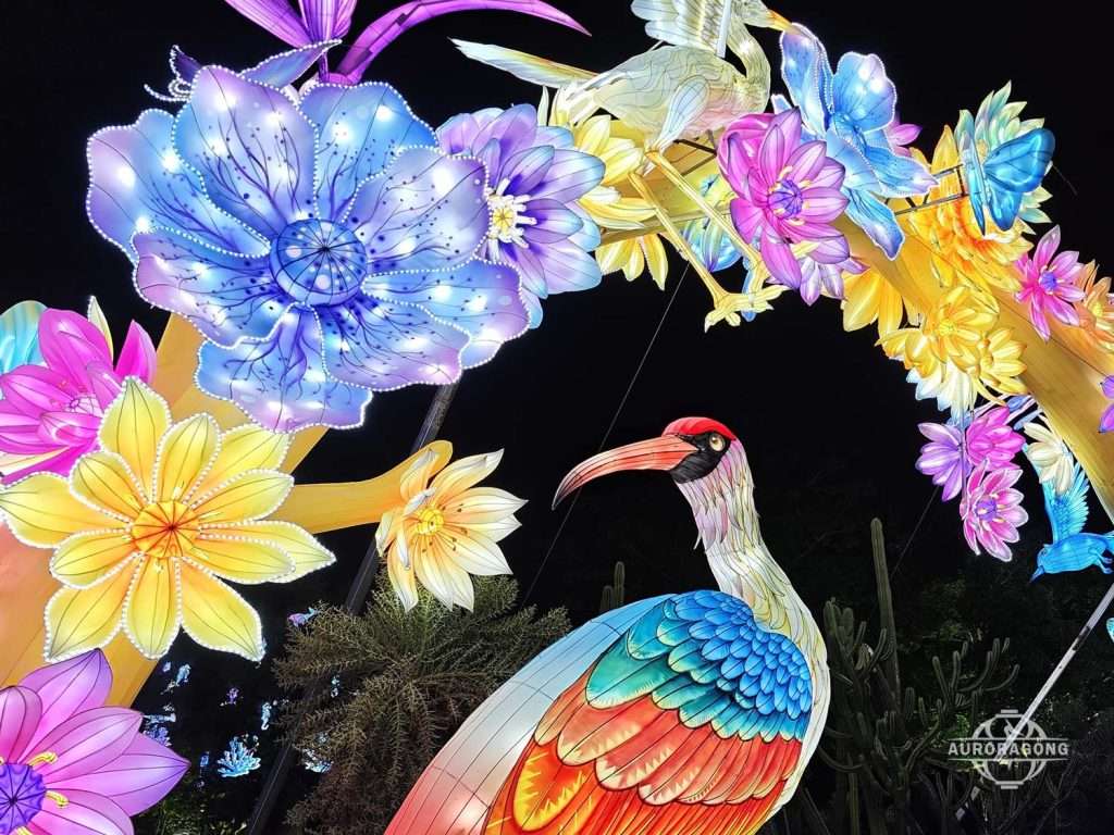 Colorful crested bird lantern with multicolored feathers among pink and yellow flowers