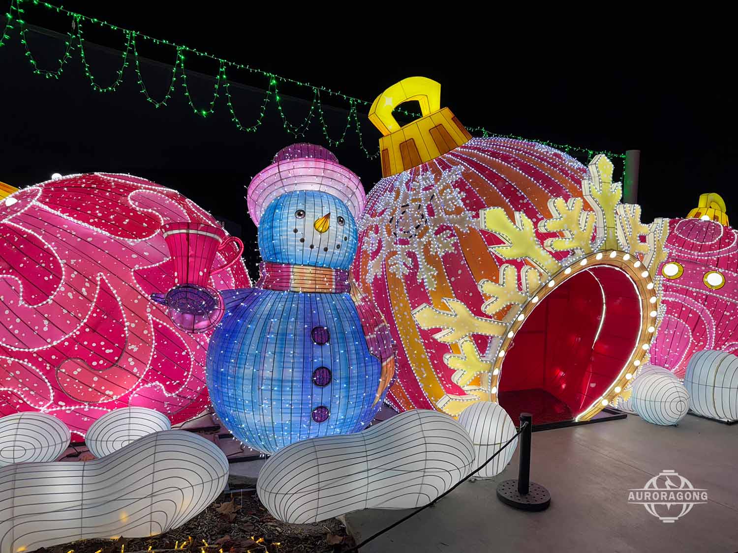 Illuminated snowman lantern with red hat beside giant pink Christmas baubles at an outdoor winter lantern festival