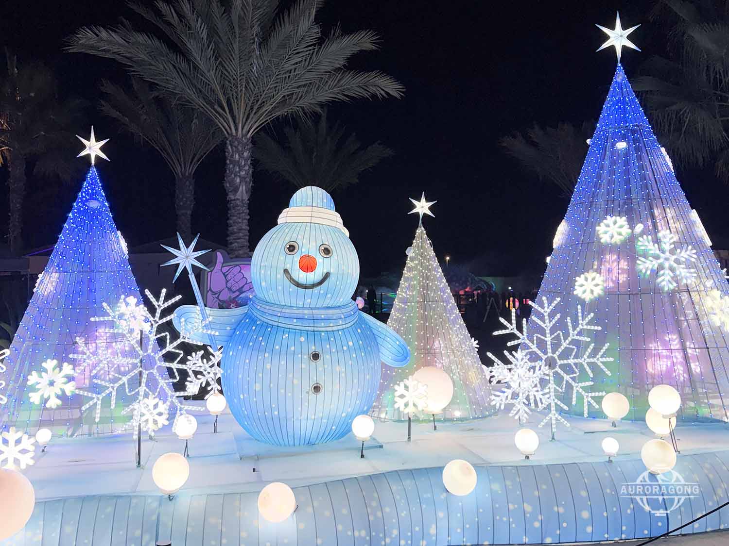Illuminated snowman lantern with snowflake wand beside blue Christmas trees at an outdoor winter lantern festival